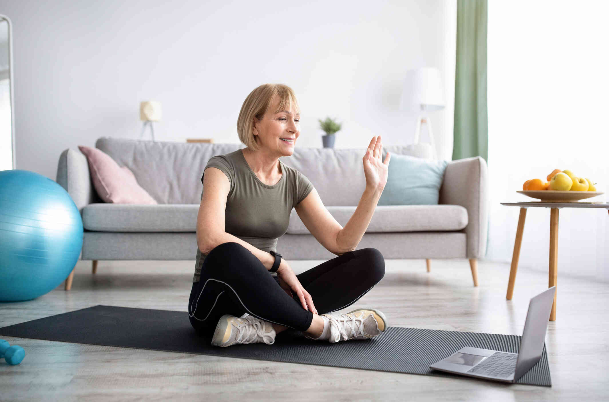 A middle aged woman sits on a yoga mat in her livingroom and waves at the laptop screen that is open on the matt infront of her with a smile during a video call.